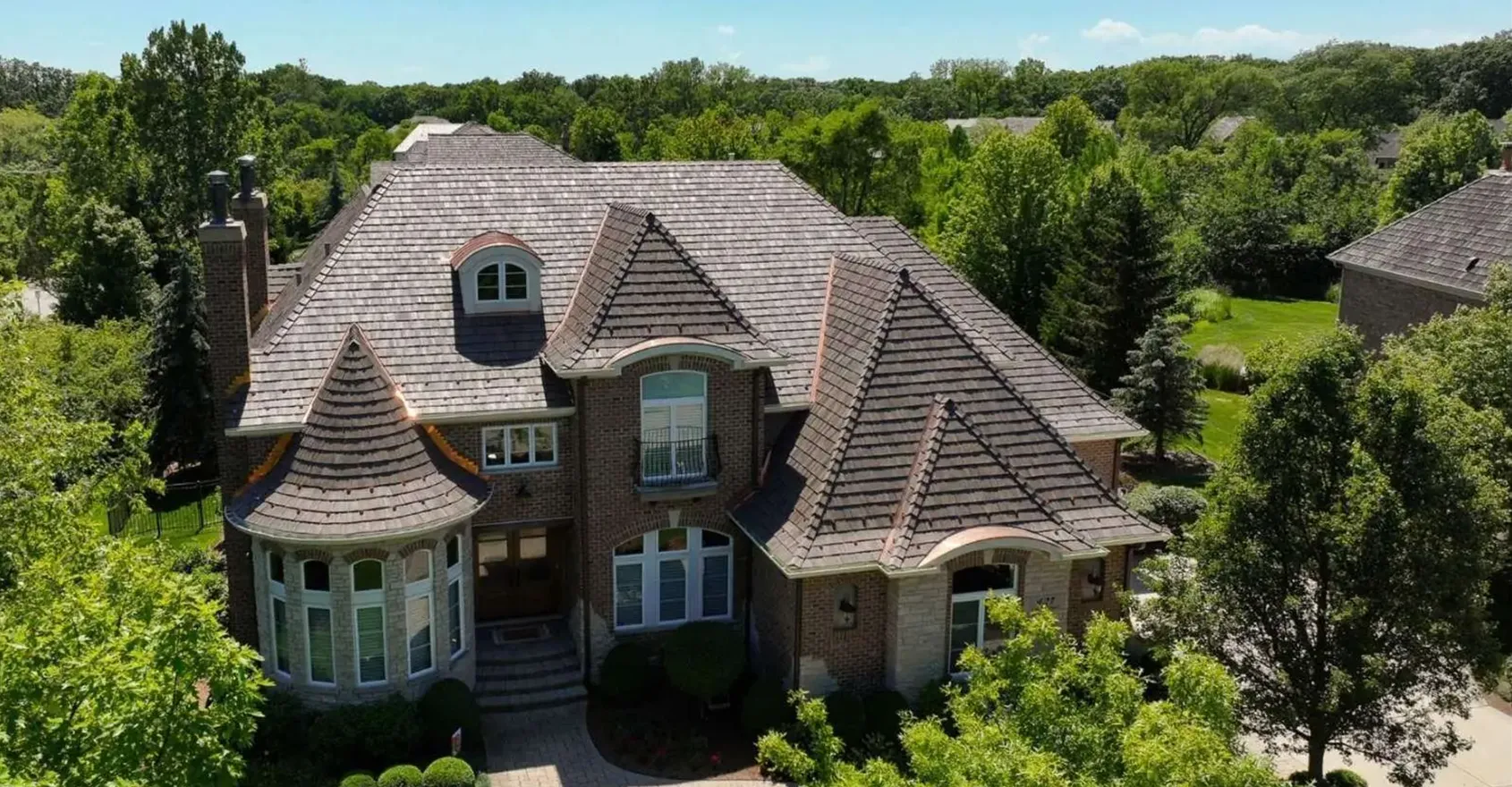 Aerial view of a large brick home with a steep, multi-gabled shingle roof and turret